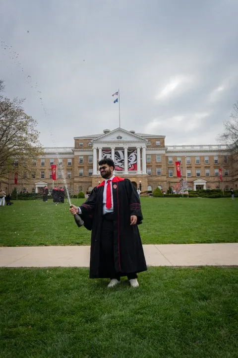 Karan popping champagne in a graduation gown in front of Bascom Hall